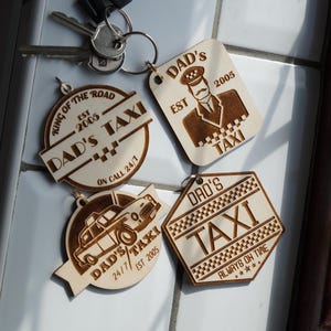 Op de afbeelding: Een verzameling houten sleutelhangers met een taxi-thema. De sleutelhangers hebben gegraveerde ontwerpen, waaronder een taxichauffeur, een taxi en de woorden "DAD'S TAXI" en "EST 2005". Sommige sleutelhangers bevatten ook de zinnen "KING OF THE ROAD" en "ALWAYS ON TIME".