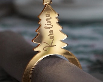 May include: A gold-colored Christmas tree-shaped napkin ring with the name "Karolina" engraved on it. The ring is wrapped around a folded brown napkin, with a star at the top of the tree.