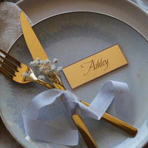 May include: A gold-colored fork and knife are arranged on a plate with a white ribbon and a name card that reads "Ashley". The plate is light gray with a textured surface. Small white flowers are tucked between the utensils.