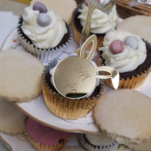 Op de afbeelding: Een etagère met cupcakes en koekjes. De cupcakes zijn bedekt met witte glazuur en gegarneerd met kleine, kleurrijke snoepjes. Een goudkleurige konijnvormige decoratie met de tekst "HAPPY Easter" staat op de voorgrond. De koekjes zijn rond en licht van kleur.