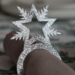 May include: A silver glitter star-shaped napkin ring. The ring is wrapped around a brown napkin. The star has a five-point design with leaf-like accents. The napkin ring is a decorative item for table settings.