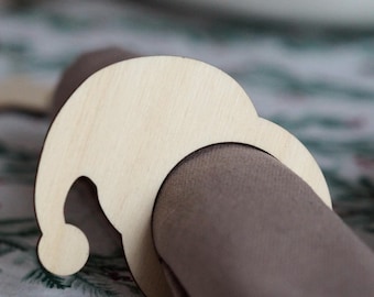 May include: A wooden Santa hat napkin ring. The ring is made of light-colored wood and is shaped like a Santa hat. The napkin ring is wrapped around a brown napkin, set on a festive tablecloth.
