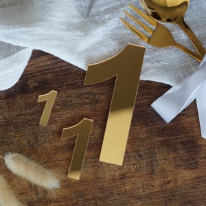 Può includere: Decorazioni dorate con il numero "1" in tre dimensioni, disposte su una superficie di legno scuro. Una forchetta e un cucchiaio dorati sono legati con un nastro bianco. Un panno bianco e fiori secchi sono anch'essi presenti, creando un tema celebrativo.