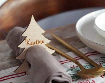 May include: A wooden Christmas tree-shaped place card holder with the name "Karolina" engraved on it, resting on a rolled-up gray napkin. Gold-colored flatware and a white bowl are in the background. The table setting is on a festive Christmas-themed tablecloth.