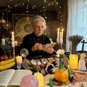 May include: An elderly person is seated at a table, reading tarot cards. The table is adorned with candles, crystals, a crystal ball, and a small pumpkin. The scene is illuminated by soft lighting, creating a mystical atmosphere.