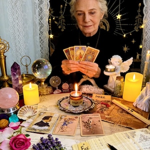 May include: A woman holding tarot cards, surrounded by candles, crystals, and a crystal ball. The scene includes a lit candle in a decorative holder, a rose, and a sprig of lavender. The background features a black cloth with gold stars.