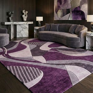 Könnte beinhalten: Ein großer rechteckiger Teppich mit einem mehrschichtigen abstrakten Muster in den Farben Dunkelviolett, Lavendel und Grau. Das Design umfasst geschwungene, fließende Linien und Bereiche mit linearen Streifenstrukturen auf der Oberfläche.