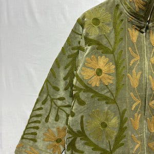 May include: Olive green velvet jacket with intricate floral embroidery in shades of gold and green. The jacket features a high collar and a full-length zipper. The floral design includes large daisy-like flowers and stylized leaves.