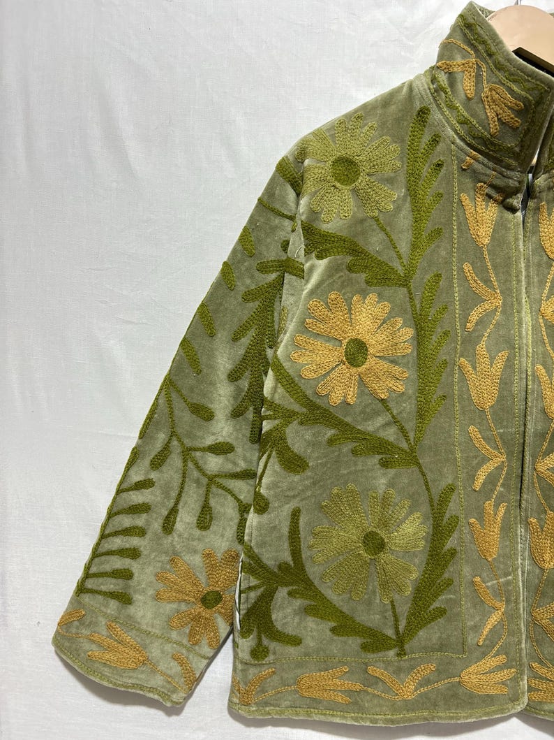 May include: Olive green velvet jacket with intricate floral embroidery in shades of gold and green. The jacket features a high collar and a full-length zipper. The floral design includes large daisy-like flowers and stylized leaves.