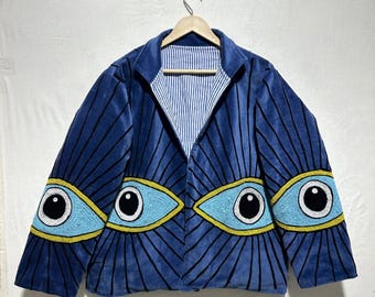 May include: A blue velvet jacket with a unique eye design. The jacket features large, beaded eye motifs in light blue, yellow, and black on the sleeves and front. The interior lining is striped. The jacket is hanging on a wooden hanger.