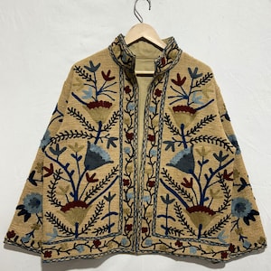 Puede incluir: Chaqueta beige con intrincados bordados florales en azul, rojo y beige. La chaqueta presenta un cuello alto y un patrón detallado de flores y hojas estilizadas. El bordado es denso, creando una superficie texturizada.