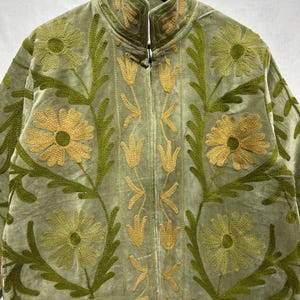 May include: A sage green velvet jacket with intricate floral embroidery in shades of green and gold. The jacket features a high collar and a zip-up front. The embroidered design includes large flowers, leaves, and stylized floral patterns, creating a decorative and textured look.