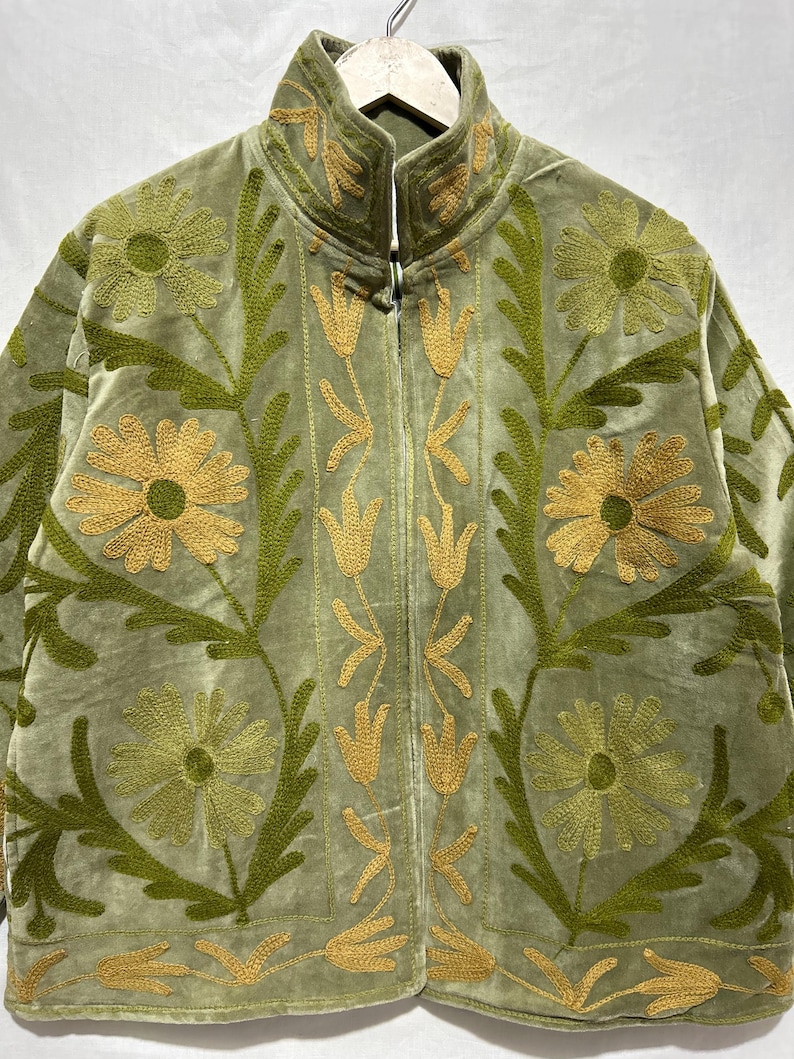 May include: A sage green velvet jacket with intricate floral embroidery in shades of green and gold. The jacket features a high collar and a zip-up front. The embroidered design includes large flowers, leaves, and stylized floral patterns, creating a decorative and textured look.