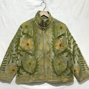 May include: A sage green jacket with a high collar and a floral embroidery design. The jacket features a zipper closure and is adorned with embroidered flowers and leaves in shades of gold and green. The jacket is hanging on a wooden hanger.