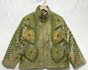 Puede incluir: Chaqueta verde salvia con cuello alto y bordado floral. La chaqueta tiene cierre de cremallera y está adornada con flores y hojas bordadas en dorado y verde. La chaqueta está colgada en una percha de madera.