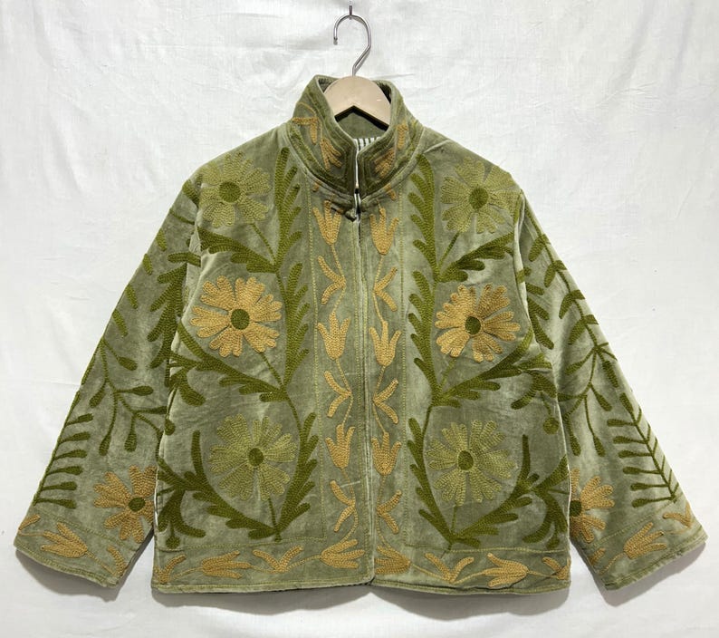May include: A sage green jacket with a high collar and a floral embroidery design. The jacket features a zipper closure and is adorned with embroidered flowers and leaves in shades of gold and green. The jacket is hanging on a wooden hanger.