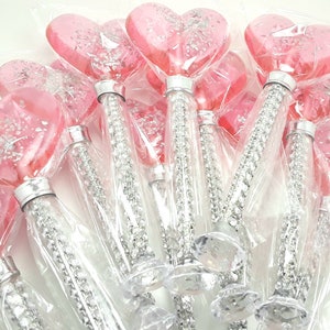 Peut inclure: Un groupe de sucettes en forme de cœur rose avec des paillettes argentées sur des bâtonnets enveloppés dans du plastique transparent avec des paillettes argentées.