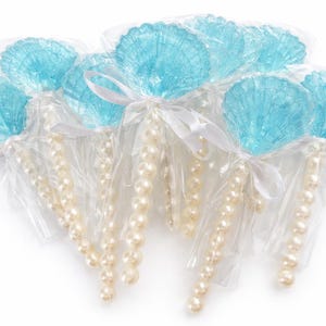 May include: A group of seashell-shaped lollipops in clear cellophane. The lollipops are a translucent blue, with white pearl bead strands and white ribbon. The candy is arranged on a white background.