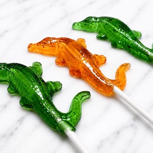 May include: Three alligator-shaped lollipops attached to white sticks. Two of the lollipops are translucent green, and one is translucent orange. Each candy features a detailed moulded texture representing the head, body, legs, and tail of an alligator.