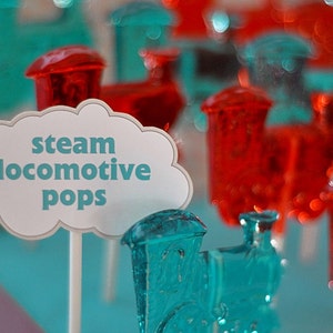 Puede incluir: Un cartel blanco en forma de nube con el texto "steam locomotive pops" en letras azules. El cartel está delante de una exposición de piruletas en forma de tren azules y rojas.