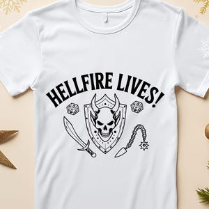 May include: White t-shirt with the text "HELLFIRE LIVES!" in black. The graphic features a skull with horns, a sword, a flail, and dice. The design is centered on the front of the shirt.