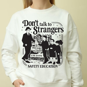 May include: A cream-coloured crewneck sweatshirt featuring a black graphic print. The design depicts a man in a suit shaking hands with a child, with two other children nearby. Text reads: Don't talk to Strangers, HI MR. WHATSI, and SAFETY EDUCATION.