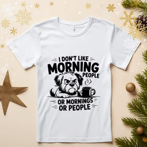 May include: White t-shirt with a black graphic of a grumpy dog and a coffee mug. The text reads "I DON'T LIKE MORNING PEOPLE OR MORNINGS OR PEOPLE."