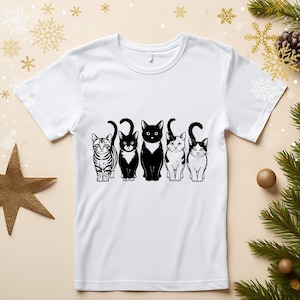 Puede incluir: Camiseta blanca con un diseño de cinco gatos en blanco y negro en fila. Los gatos tienen diferentes marcas, incluyendo rayas y patrones de esmoquin. El diseño es una ilustración gráfica sencilla.