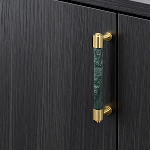 May include: Cabinet handles with a gold finish and a dark green, textured central section. The handles are attached to a dark wood cabinet with visible grain. The cabinet is topped with a dark grey countertop.