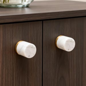 Könnte beinhalten: Zwei weiße Marmor-Schrankknöpfe mit goldfarbenen Basen sind an einem dunkelbraunen Holzschrank befestigt. Die Knöpfe sind zylindrisch und haben eine polierte Oberfläche. Eine Glasvase mit Blumen steht auf dem Schrank.