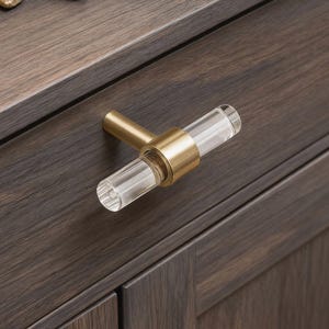 May include: A clear acrylic and brass cabinet pull. The cylindrical acrylic handle is held in place by a brass ring and stem. The pull is mounted on a dark brown wooden cabinet door.