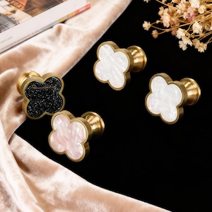 May include: Four decorative cabinet knobs with a cloverleaf design. The knobs have a brass base and feature cloverleaf faces in black, white, and pink. The knobs are arranged on a beige and black background.