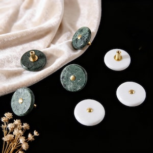 Puede incluir: Una colección de pomos redondos para armarios. Algunos son de mármol verde con herrajes dorados, otros de mármol blanco con herrajes dorados. Los pomos están dispuestos sobre una superficie negra con una tela color crema en el fondo.