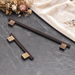 Könnte beinhalten: Zwei dunkelbraune Schrankgriffe mit einem strukturierten, zylindrischen Griff und quadratischen, messingfarbenen Endstücken. Ein Griff ist länger als der andere. Die Griffe werden auf einer dunkelgrauen Oberfläche präsentiert.