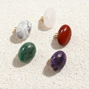 May include: Five gemstone cabinet knobs in a circular arrangement. The knobs are oval-shaped and feature a variety of colors and patterns, including white marble, clear quartz, red jasper, green aventurine, and purple amethyst. Each knob has a gold-colored base.