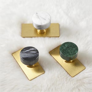 Może przedstawiać: Trzy dekoracyjne gałki do szafek z marmurowymi blatami w kolorze białym, czarnym i zielonym. Każda gałka jest zamontowana na prostokątnej podstawie z mosiądzu. Gałki są prezentowane na białej, teksturowanej powierzchni.