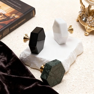 May include: A collection of decorative cabinet knobs in black, white, and green marble. Each knob has a faceted, geometric design and is mounted on a gold-toned base. The knobs are displayed on a white, textured surface.