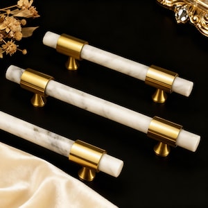 May include: Three cabinet pulls with white marble centers and gold-toned metal accents. The pulls have a cylindrical design with a gold band at each end and a small, decorative gold base. The marble has gray veining, and the pulls are arranged on a black surface.