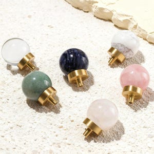 May include: A selection of decorative cabinet knobs with spherical tops in various colours and materials. The knobs feature clear, white marble, blue, green, and pink spheres, each mounted on a brass base. These knobs are designed for furniture and home decor.