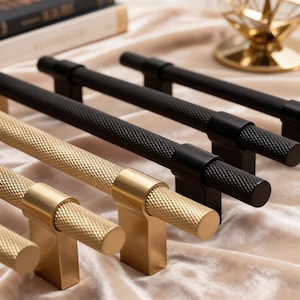May include: A selection of cabinet pulls in gold and black. The gold pulls have a textured, cylindrical handle and a square base. The black pulls have a similar textured handle and a rectangular base. The pulls are arranged in a row.