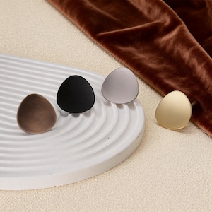 Op de afbeelding: Vier decoratieve kastknoppen in brons, zwart, grijs en goud. Elke knop heeft een gladde, afgeronde driehoekige vorm. De knoppen worden gepresenteerd op een wit, geribbeld oppervlak met een bruine fluwelen stof op de achtergrond.