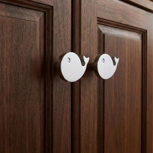 May include: Two polished chrome whale-shaped cabinet knobs mounted on a dark brown wooden cabinet door. The knobs have a smooth, reflective surface and a small, circular eye detail. The cabinet door has vertical wood grain.