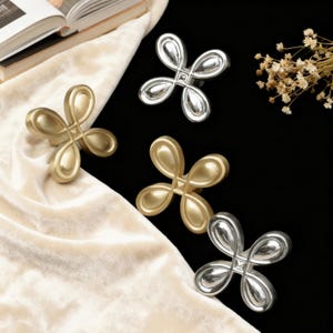 May include: Four decorative cabinet knobs in a cloverleaf design. Two are gold-toned, and two are silver-toned. The knobs are arranged on a black and cream-coloured surface, with a book and dried flowers in the background.