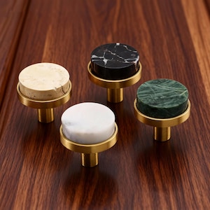 Op de afbeelding: Vier kastknoppen met ronde marmeren bladen in beige, zwart, wit en groen. Elke knop heeft een goudkleurige basis en een ronde goudkleurige ring. De knoppen worden gepresenteerd op een donker houten oppervlak.