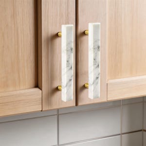 Puede incluir: Dos puertas de armario con herrajes rectangulares de mármol y latón. El mármol es blanco con vetas grises. Los herrajes de latón son redondos. Las puertas del armario son de madera clara.