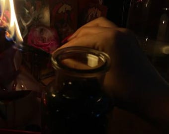 May include: A dark glass jar filled with a dark liquid, possibly a potion or herbal infusion. A hand with red nail polish is near the jar. A flame is lit nearby, casting shadows and creating a mystical ambiance. The scene suggests a ritual or spiritual practice.