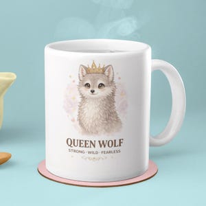 May include: White ceramic mug featuring a watercolor illustration of a wolf wearing a gold crown. The text "QUEEN WOLF" is below the wolf, with "STRONG WILD FEARLESS" underneath. The mug sits on a pink coaster.