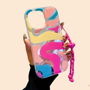 May include: Clear phone case with a colorful abstract design in pink, yellow, blue, and peach. The case has a pink heart-shaped charm and a matching beaded wrist strap. The case is designed to fit a smartphone.