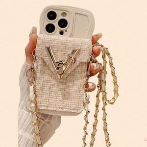 Puede incluir: Funda de teléfono beige con textura tejida y correa de cadena dorada. La funda presenta un emblema dorado en forma de V y un pequeño elemento decorativo. La funda está diseñada para guardar un smartphone.
