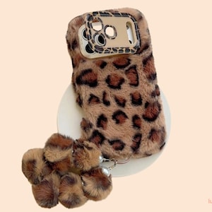 May include: A fluffy phone case with a leopard print design in shades of brown and black. The case has a matching pom-pom charm with a pearl accent. The phone case is designed to protect the phone from scratches and damage.